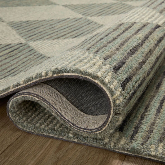 A close-up of a folded area rug showing its textured woven pattern in muted gray, sage green, and cream tones, resting on a light wood floor.