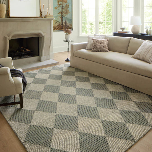 A large woven area rug with a gray and beige diamond pattern, styled in a bright living room next to a beige sofa and fireplace.